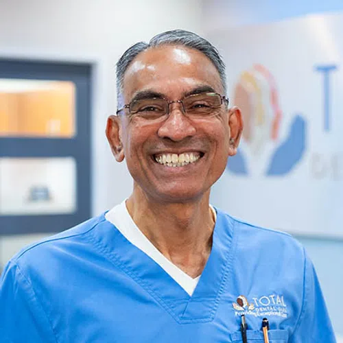 Smiling male dentist in blue scrubs and glasses in a dental clinic setting, showing friendly professionalism.