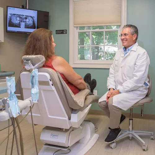 Dentist in white coat consulting with female patient in dental chair with tooth X-ray on screen.