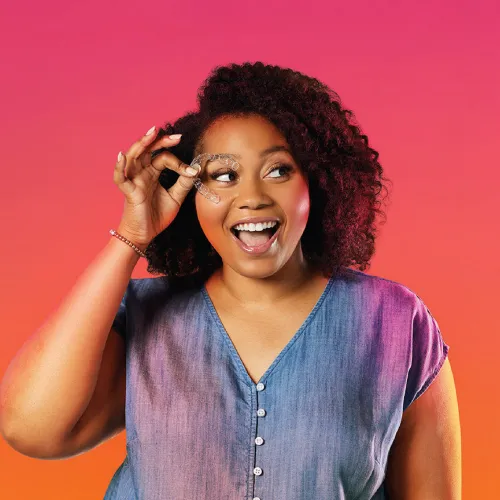 Smiling woman with curly hair wearing denim shirt poses against vibrant pink and orange gradient background.