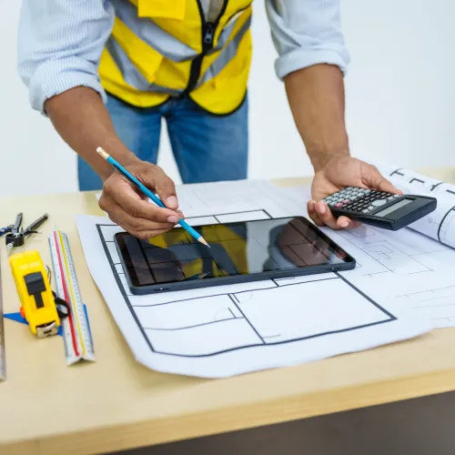 Architect in yellow vest working on blueprints with tablet, calculator, and measuring tools on wooden table.