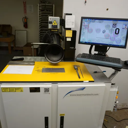 Industrial laser marking machine with computer control and measuring tools on a yellow work surface in workshop.