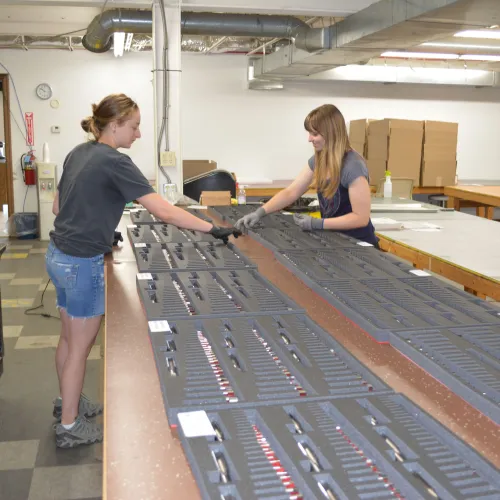 Two women assembling or inspecting organized trays of small items on a large industrial table in a warehouse.