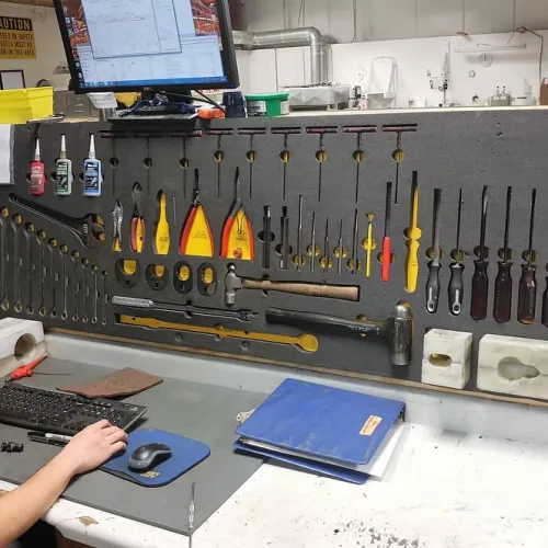 Man working at a workshop desk with organized tools hanging on a pegboard and a computer monitor above.