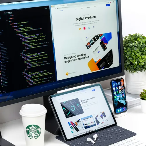 Workspace with computer monitor showing code and digital products website, tablet with app concept, smartphone, and Starbucks cup.