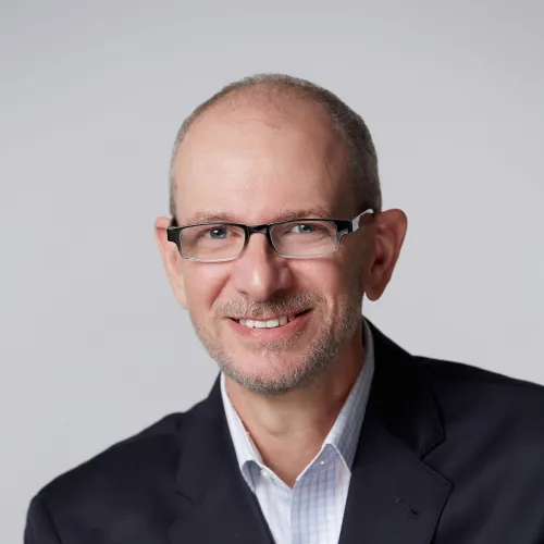 Smiling middle-aged man with glasses wearing a dark blazer and light checkered shirt against gray background