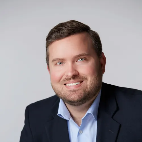 Smiling man with beard wearing blue shirt and dark blazer against light gray background