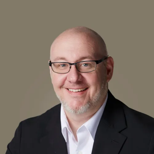 Smiling bald man with glasses in a black suit jacket and white shirt against a neutral background