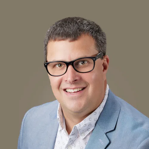 Smiling man wearing black glasses, a light blue blazer, and a white patterned shirt against a neutral background