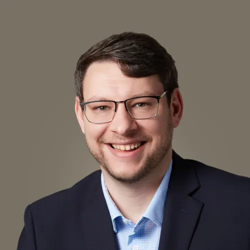 Smiling man with glasses wearing a navy blazer and light blue shirt against a beige background