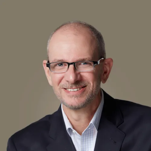 Smiling middle-aged man with glasses wearing a dark blazer and light checkered shirt against neutral background