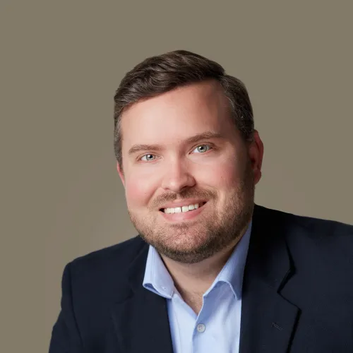 Smiling man with short brown hair and beard wearing a navy blazer and light blue button-up shirt against plain background