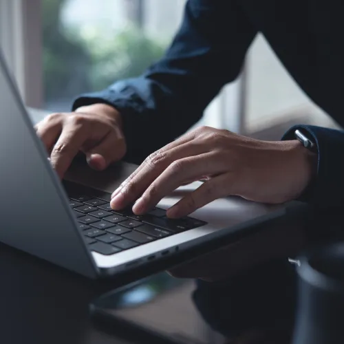 Hands typing on a laptop keyboard with a dark sleeve in a bright indoor workspace.