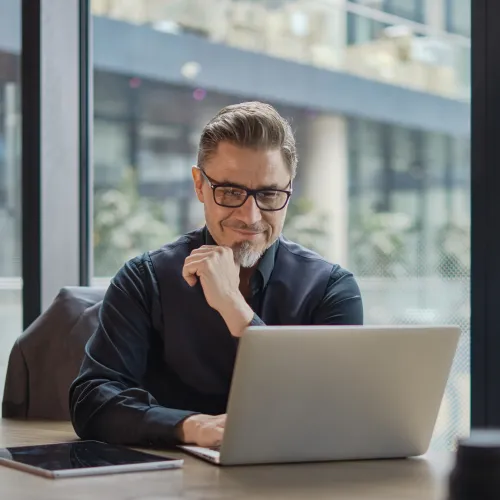 Middle-aged man wearing glasses working on laptop at modern office desk with large windows
