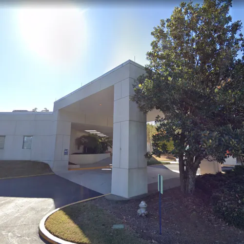 Modern white concrete building entrance with covered drive, sunlight, and a large tree to the right.
