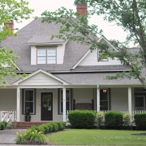Charming suburban house with a wraparound porch and lush greenery in front.