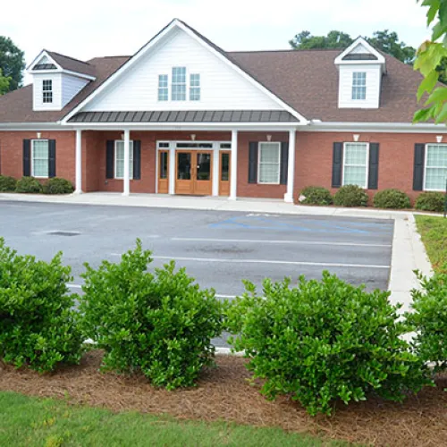 A modern brick building with a manicured lawn and parking area, surrounded by greenery.