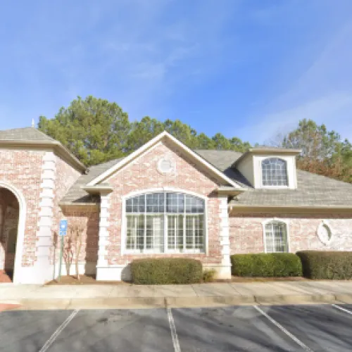 Brick building with large windows surrounded by trees and parked cars on a sunny day.