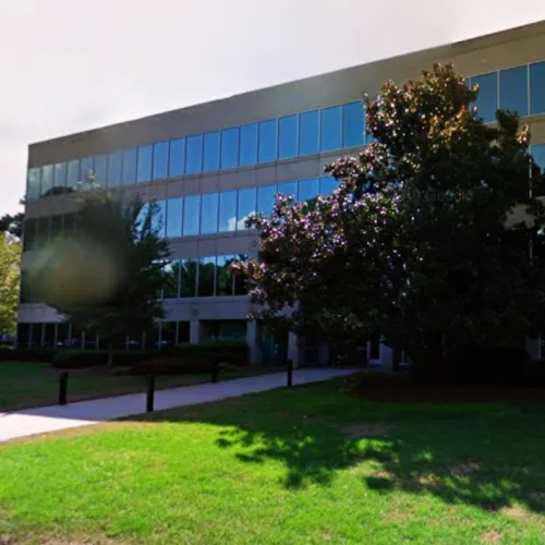 Modern office building with reflective windows, trees, and a green lawn in the foreground under clear skies