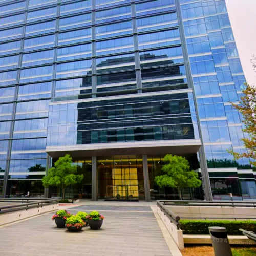 Modern glass office building with reflective windows, entrance walkway, potted plants, and green trees.