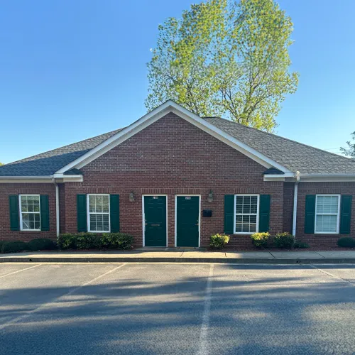 Brick duplex with green doors and shutters in a sunny suburban setting with trees and clear sky.