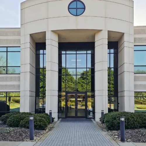 Modern office building entrance with glass doors, symmetrical windows, and landscaped walkway under clear sky