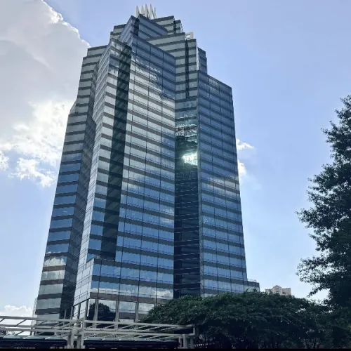 Modern glass skyscraper reflecting the sky and clouds with surrounding trees and blue sky background.