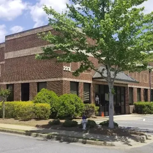 Modern brick office building with multiple windows, surrounding greenery, a tree, and a clear blue sky above.