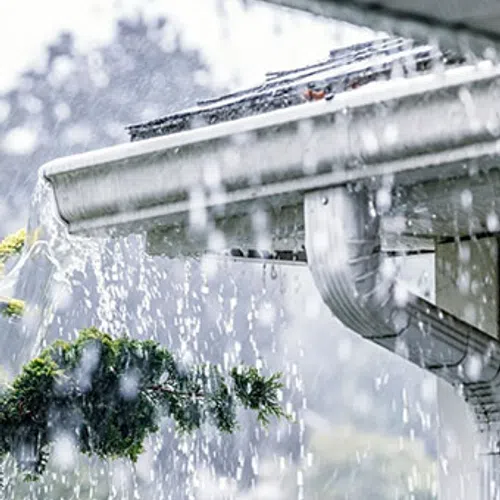 Heavy rainwater overflowing from a clogged roof gutter with water cascading down near green tree branches.