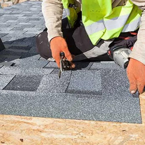 Worker wearing orange gloves and a safety vest installing gray asphalt shingles on a wooden roof deck.