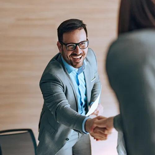 Smiling businessman in gray suit shaking hands with a colleague in a modern office setting.