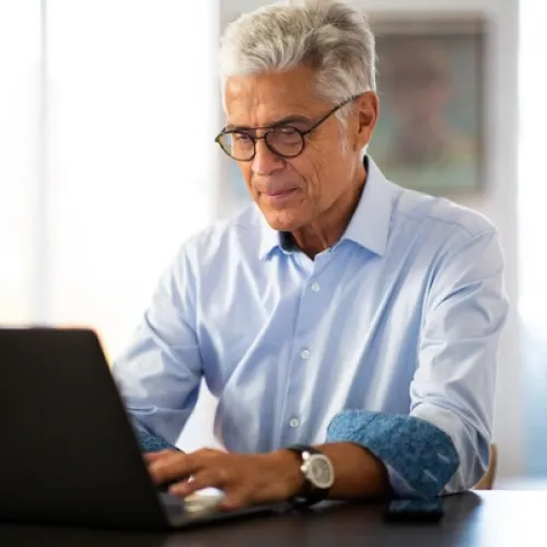 Senior man wearing glasses and blue shirt working on a laptop at a desk in a bright room