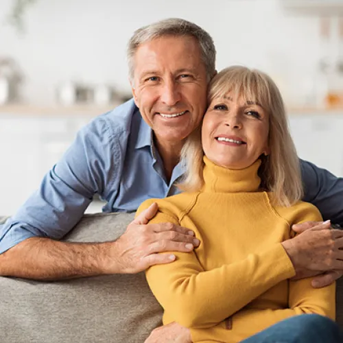 Smiling mature couple hugging on a couch in a cozy, modern living room with soft natural light.