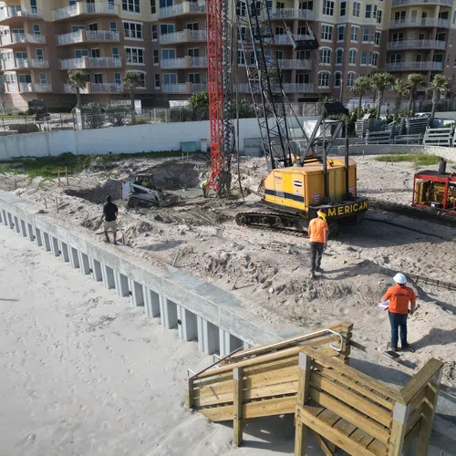 Construction site by the beach with workers, heavy machinery, sand, and a multi-story building in the background.