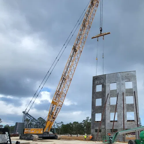 Large crane lifting a concrete wall panel with window openings at a construction site under cloudy sky.