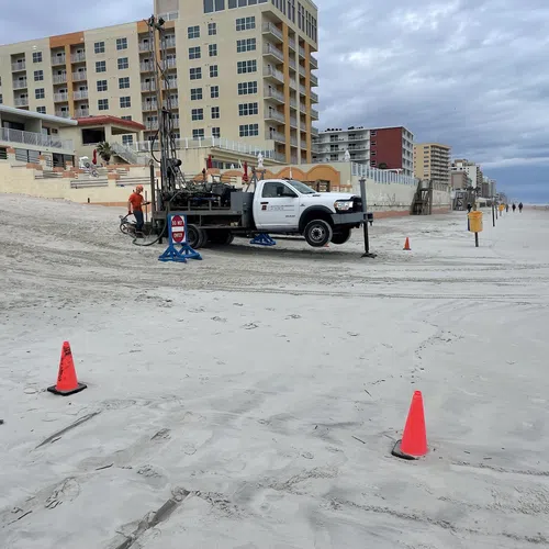 Construction truck and workers on sandy beach near orange safety cones and beachfront buildings under cloudy sky.