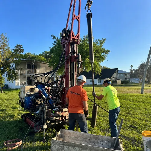 Two workers operating drilling equipment outdoors with green grass, houses, and clear blue sky in the background.
