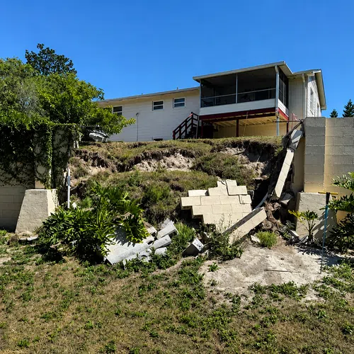 Collapsed retaining wall near a backyard with visible erosion under a residential house on a sunny day