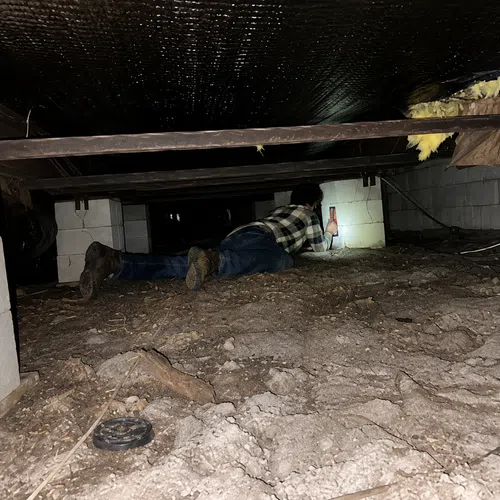 Person crawling in a dark crawl space inspecting foundation blocks using a flashlight.