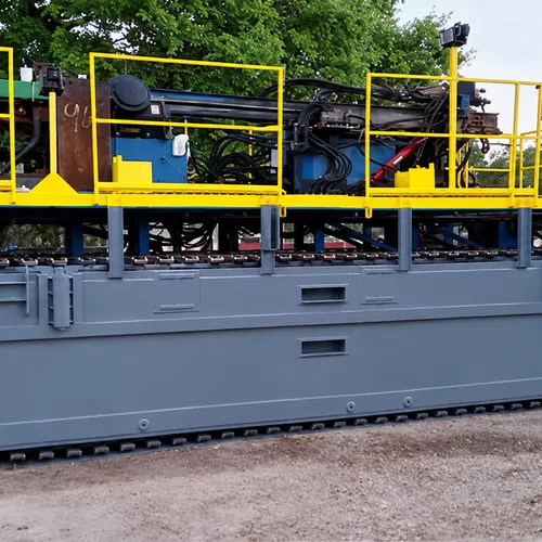 Large industrial tracked vehicle with yellow railings and complex machinery on top, set outdoors on dirt ground.