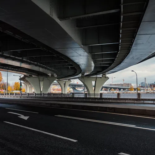 Empty road under a large modern bridge with supporting pillars and cityscape in the background under cloudy sky