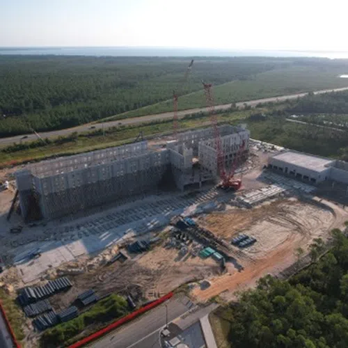 Aerial view of a large building under construction surrounded by forest and roads on a sunny day.