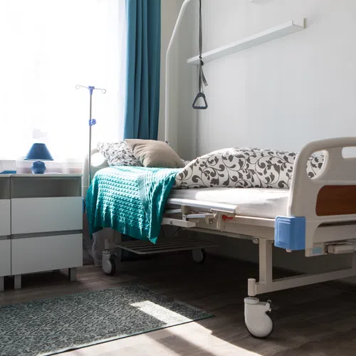 Bright hospital room with adjustable bed, teal blanket, IV stand, and dresser with lamps by the window.