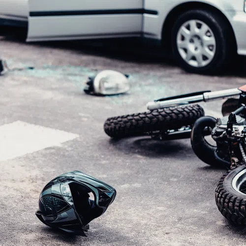 Fallen motorcycle and helmet on the road after a collision with a car in the background and shattered glass nearby.
