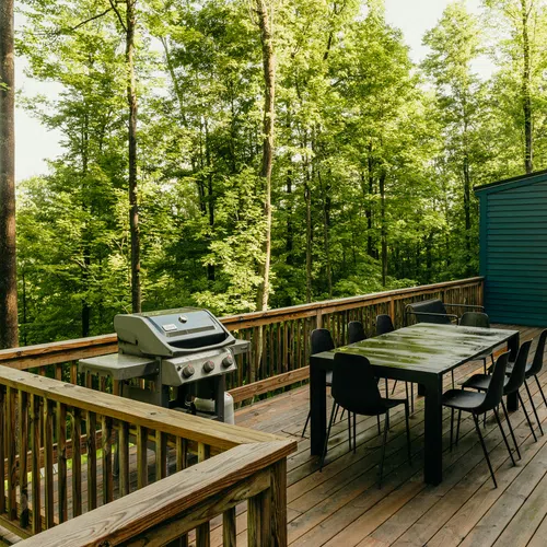 Spacious wooden deck with outdoor dining table, chairs, and a grill surrounded by lush green forest trees.