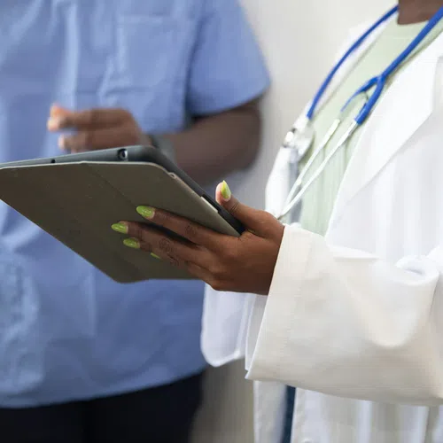 Doctor in white coat holding tablet with nurse in scrubs in background discussing patient care