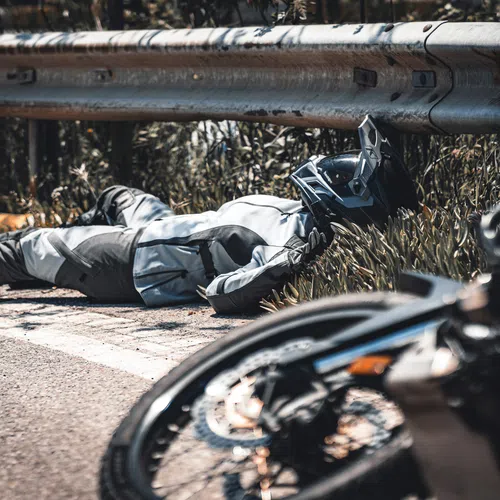Motorcyclist lying injured on roadside under guardrail with motorcycle in foreground after crash.