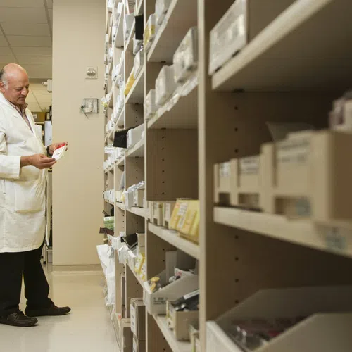 Man in white lab coat checking medication boxes in a well-organized pharmacy storage room.
