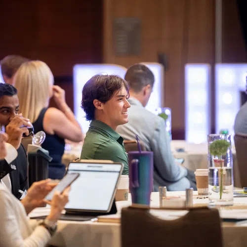 Attendees listen and take notes during a conference in a modern meeting room with round tables and drinks.