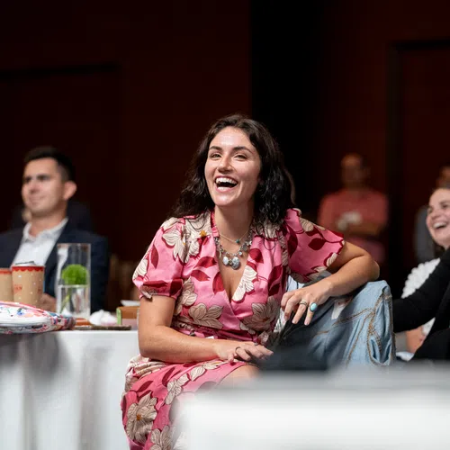 Smiling woman in pink floral dress enjoying a social event with others seated around a table indoors.