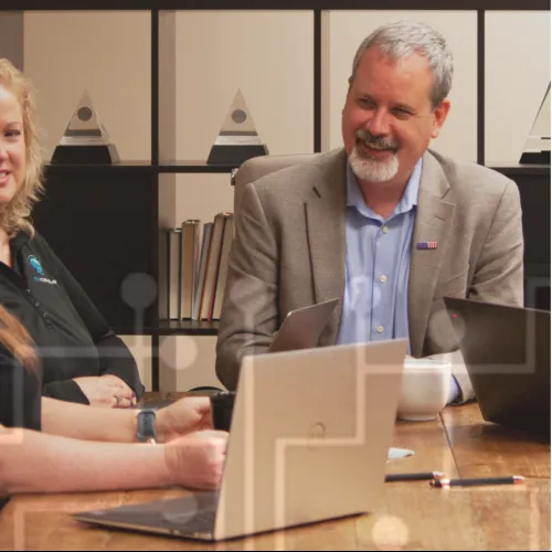 Three professionals engaged in a meeting with laptops and books in a modern office setting, smiling and discussing.